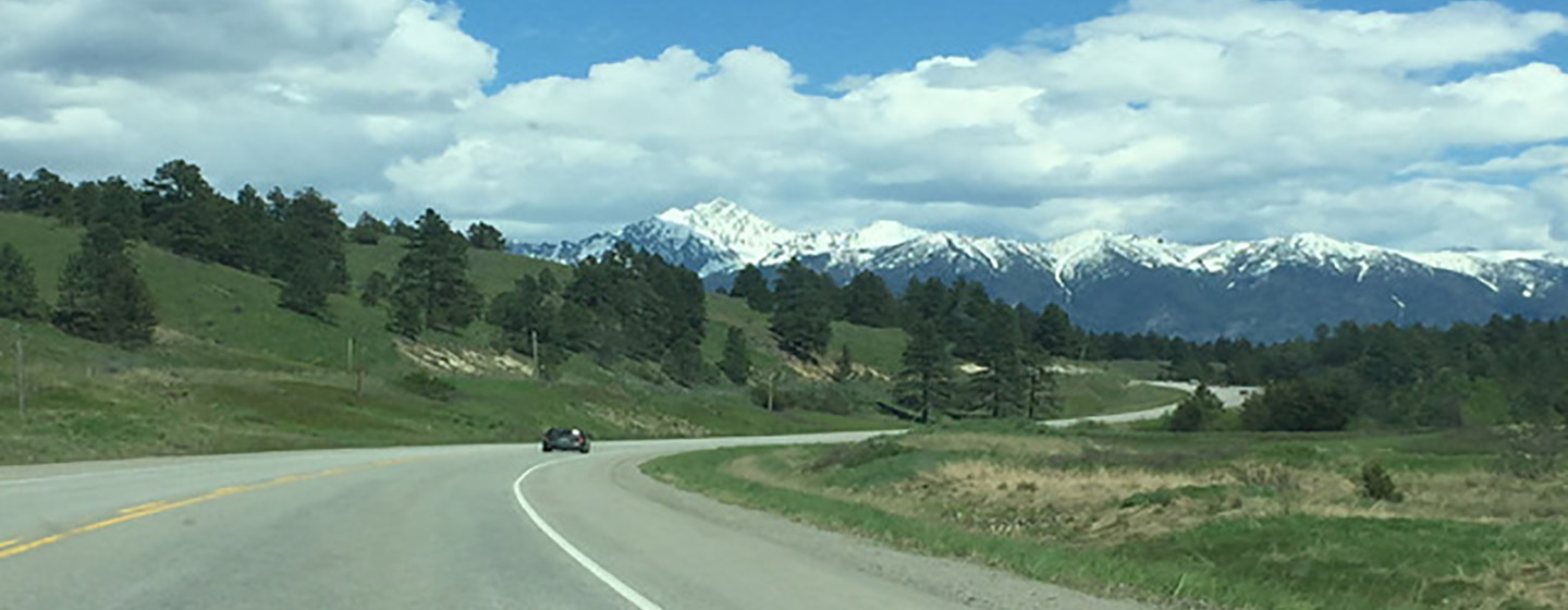 A highway winding through green foothills toward snow-capped mountains.
